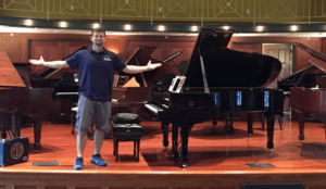 mover standing next to grand piano with arms outstretched to either side.